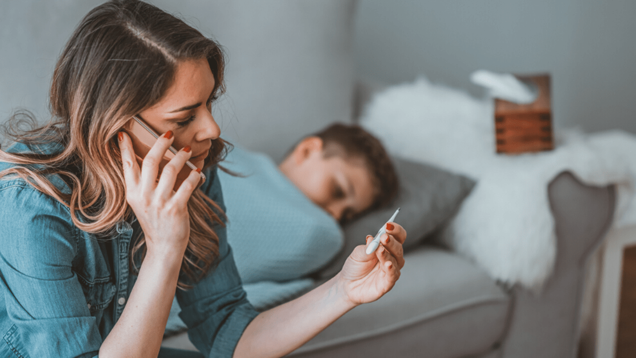 White woman on phone holding thermometer sitting on bed next to sick boy under covers