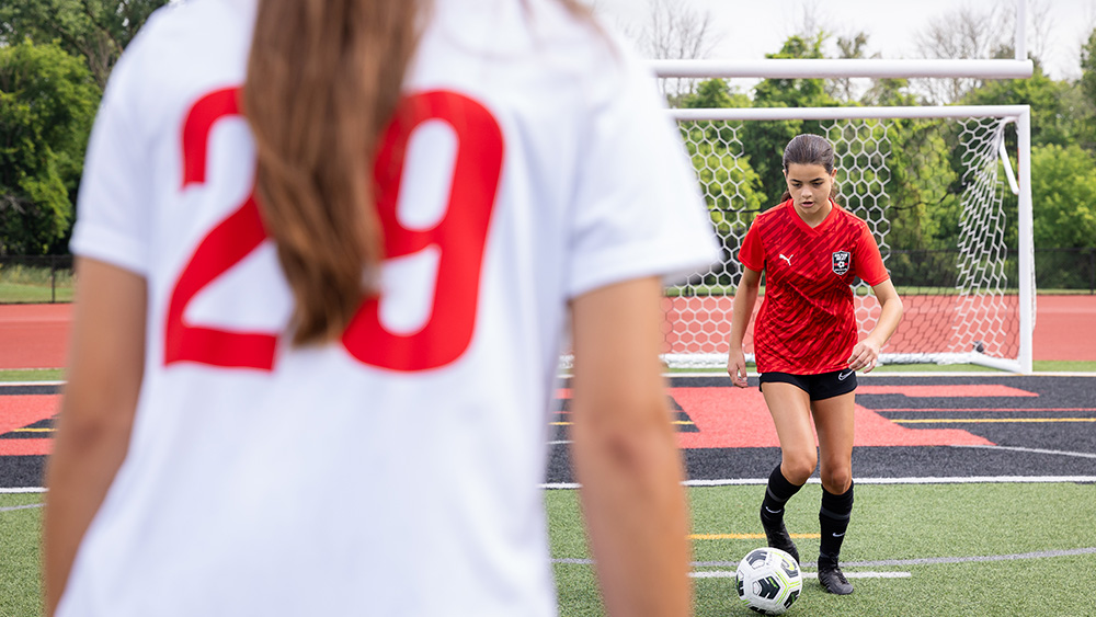 female teenage soccer player in red jersey with ball on field playing against female teenage soccer player in white jersey