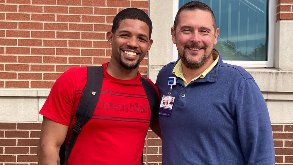 Young Black man in red shirt smiles standing next to middle aged white man in blue shirt