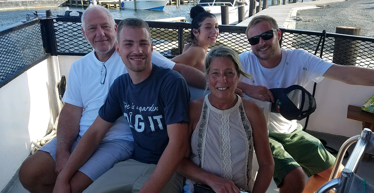 A family with two older parents and four adult children sits on a patio overlooking water. - all smiling