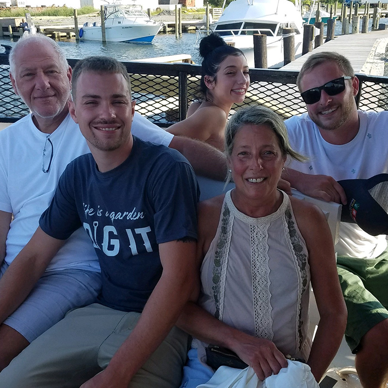 A family with two older parents and four adult children sits on a patio overlooking water. - all smiling