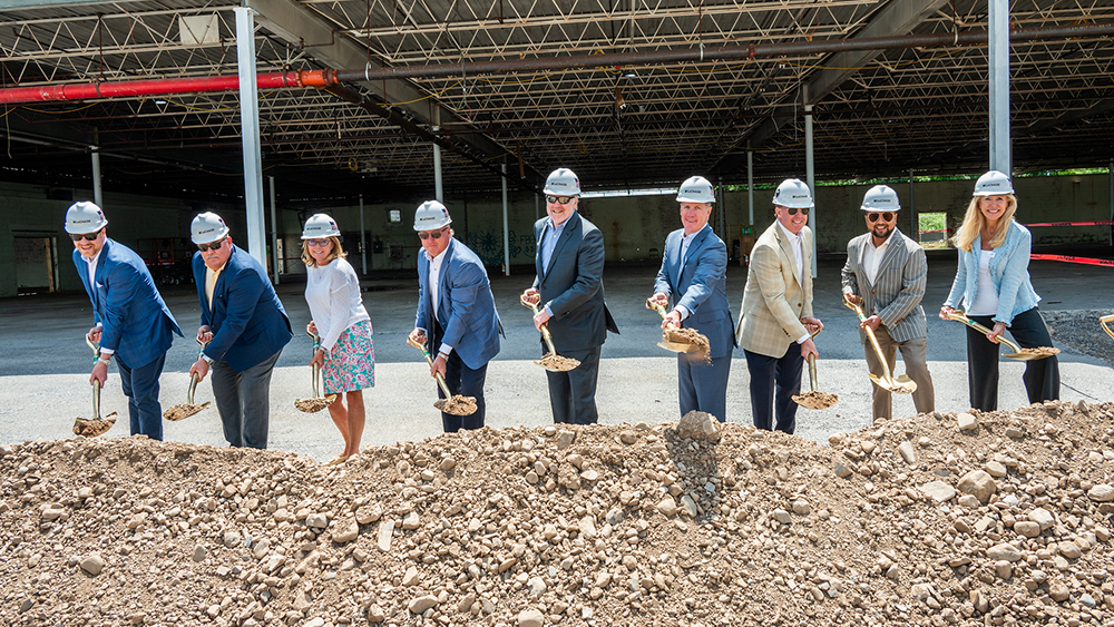 Leaders in business casual attire wearing white hard hats toss dirt using ceremonial shovels at a construction site