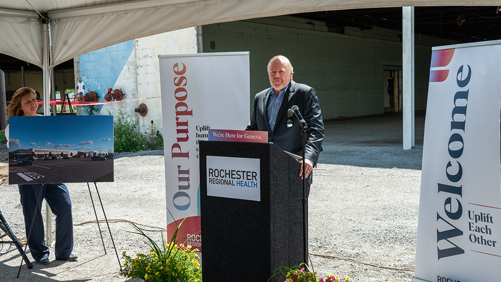 White man with white hair in business suit talks to crowd while standing next to rendering of new building