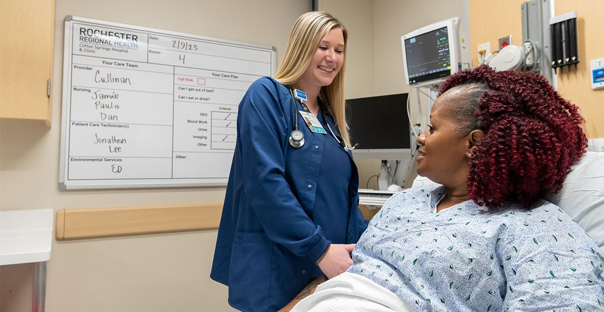 A white nurse in scrubs smiles at a smiling Black patient sitting up in a hospital bed