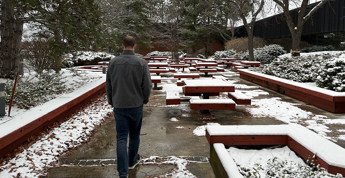 Man in winter jacket walks alone with his head down past snowy picnic tables on his right and pine trees on his left