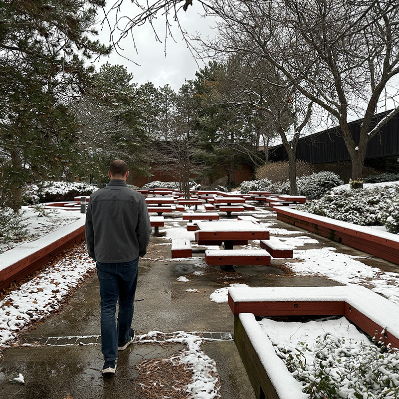 Man in winter jacket walks alone with his head down past snowy picnic tables on his right and pine trees on his left