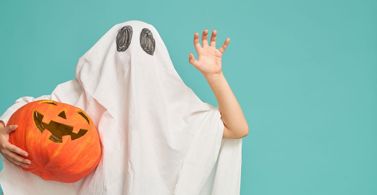 A child in a homemade ghost Halloween costume waves while holding a pumpkin in front of a teal background