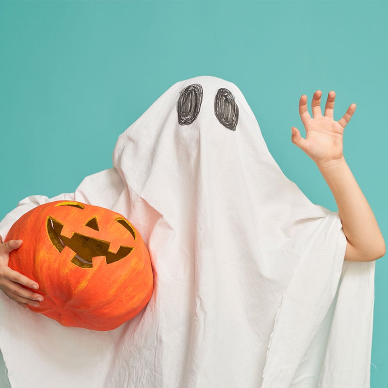 A child in a homemade ghost Halloween costume waves while holding a pumpkin in front of a teal background
