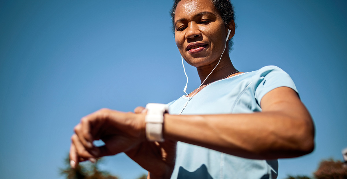 A Black woman looks at her smartwatch after exercising outside with headphones in