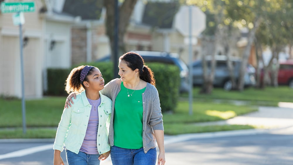 A mature Hispanic woman, in her 40s, walking with her mixed race Hispanic and African-American 11 year old daughter outdoors in their residential neighborhood. Her arm is around her the girl's shoulders as they cross a street. They are looking at each other with serious expressions.