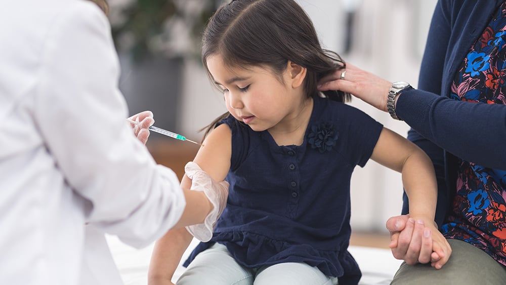 A cute young girl sits on the exam table and smiles uneasily as the nurse holds a needle in her hand and prepares to give her a vaccination shot. Her mom is holding her hand.