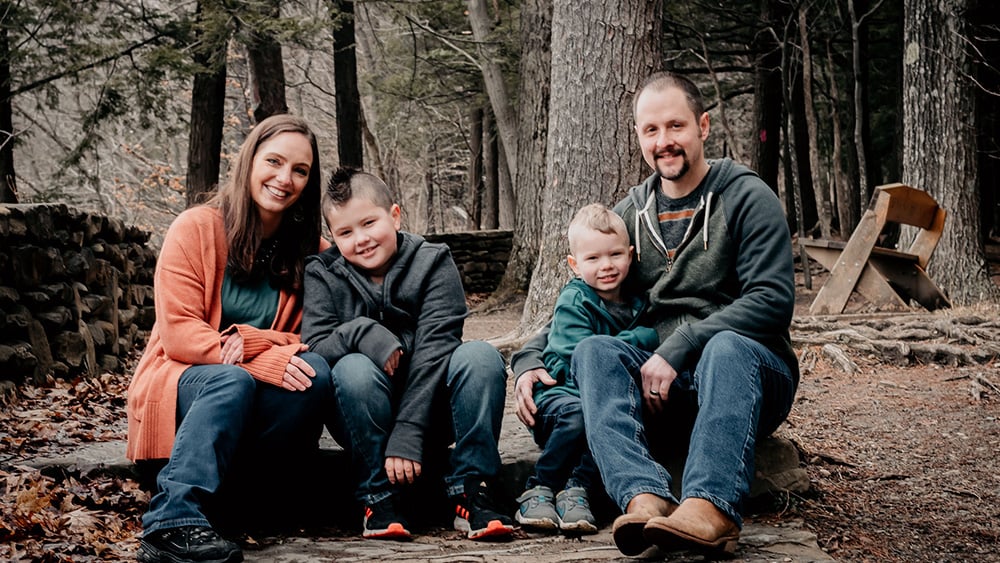 Young white family of woman man and two young boys sitting in woods outside smiling