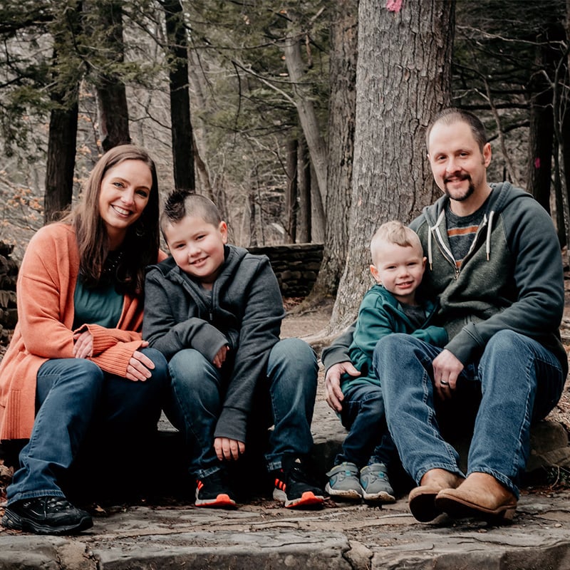 Young white family of woman man and two young boys sitting in woods outside smiling