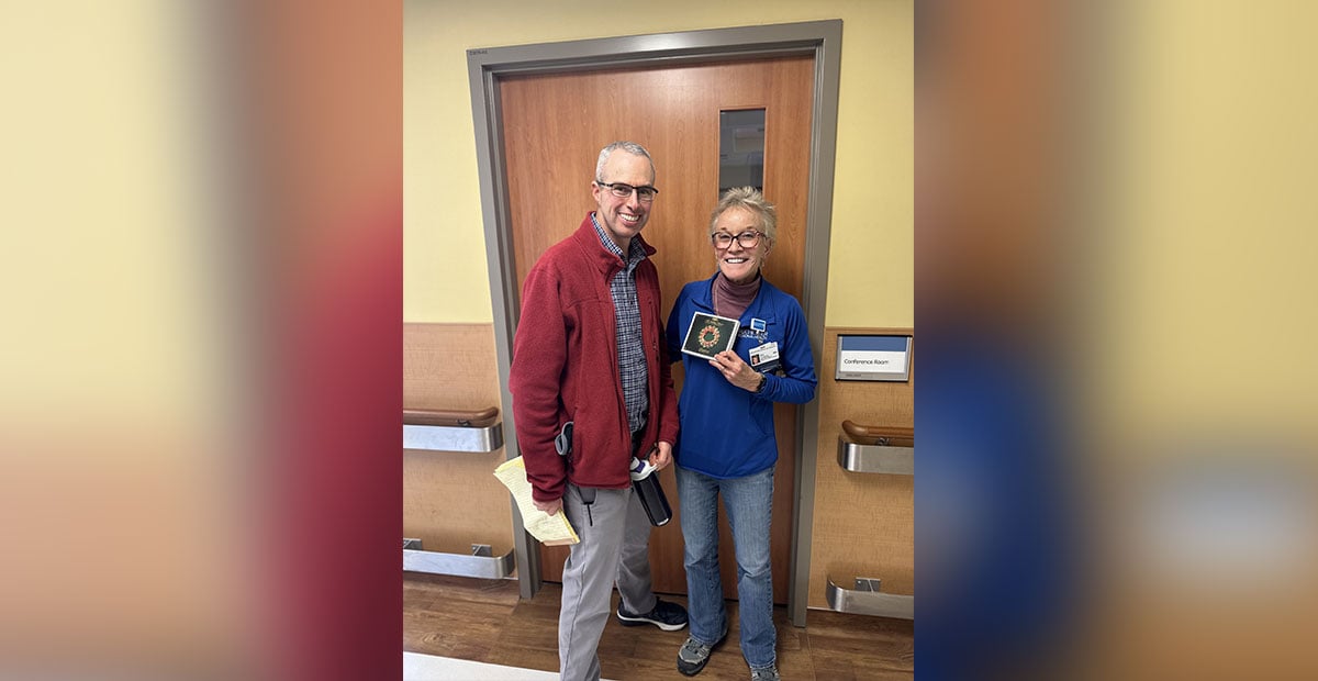 A white man wearing glasses and dressed in a maroon jacket stands next to a white female doctor wearing a blue sweater. Both are smiling. The doctor is holding a Christmas music CD.