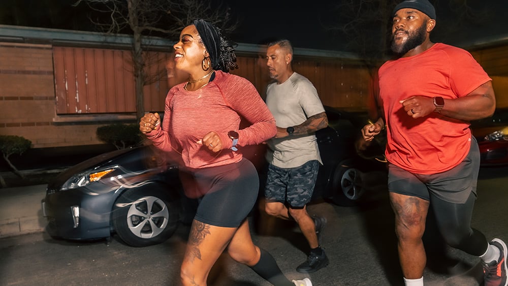 A blurred action shot of a group of friends, one Hispanic man and two Black people, jogging to the left while on a nighttime run in Los Angeles.