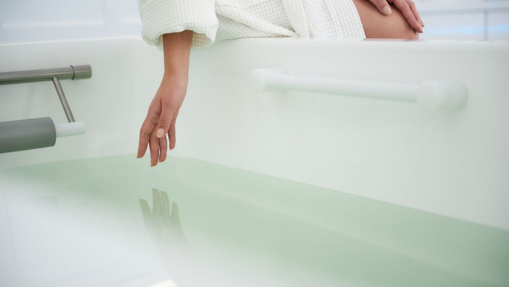 Mineral bath at spa with person in robe placing fingers into water to test heat