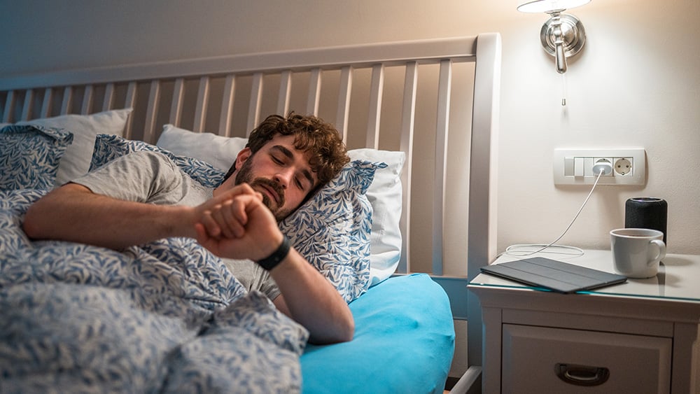 A restless Caucasian male lies in bed, gazing at his wristwatch indicating sleeplessness. Bedroom setting with a laptop and a cup nearby, reflecting common sleep disorder scenarios.
