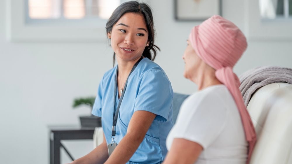 A compassionate nurse in blue uniform provides emotional support to a cancer patient wearing a pink headscarf, fostering trust and understanding in a home setting with a modern and calming interior