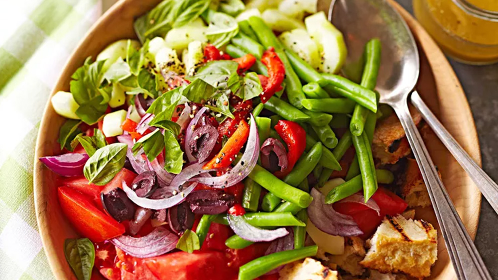 Green beans bell pepper red onion and other garden vegetables over toasted bread chunks in large wooden salad bowl