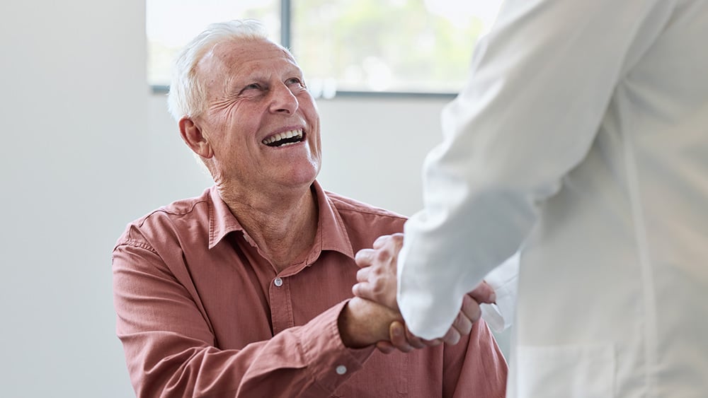 Smiling senior male holding hand of doctor. Happy elderly man talking with healthcare expert. They are doing handshake.