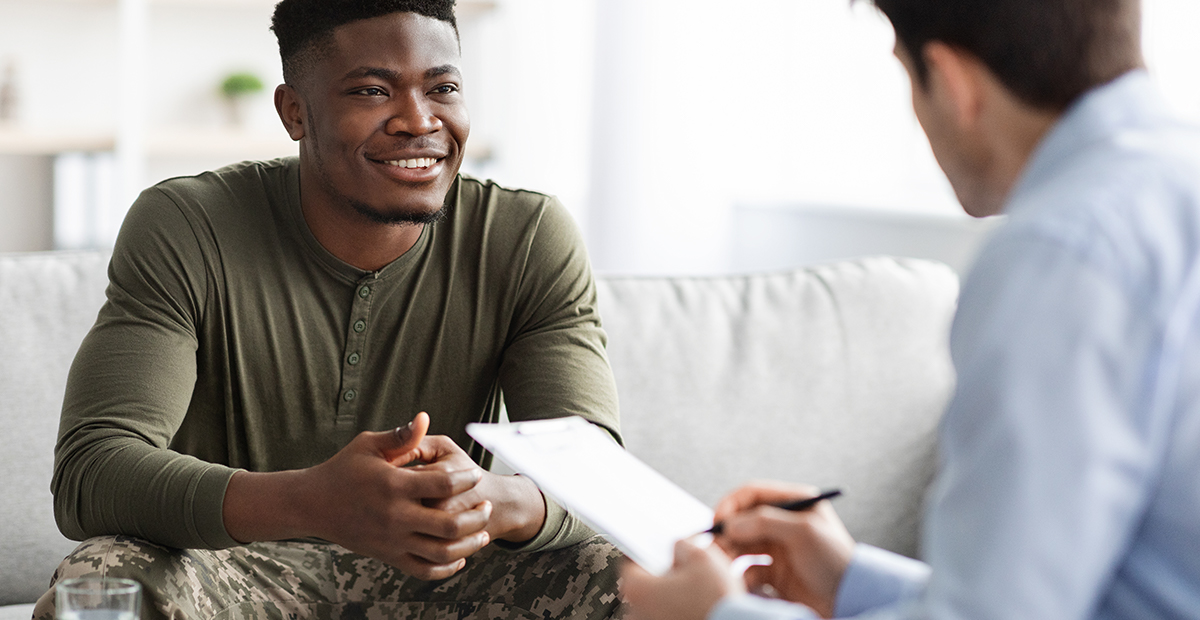 Happy young black military man in camouflage uniform sitting on couch, listening to his psychologist and smiling, enjoying results of psychotherapy