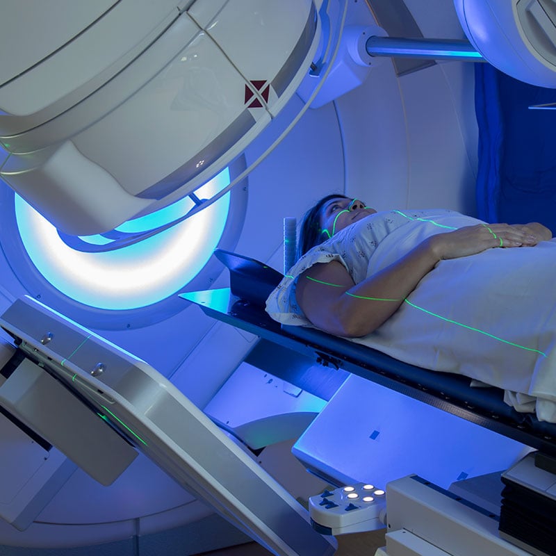 A patient in a gown lies on a table as a radiation therapy machine moves around her