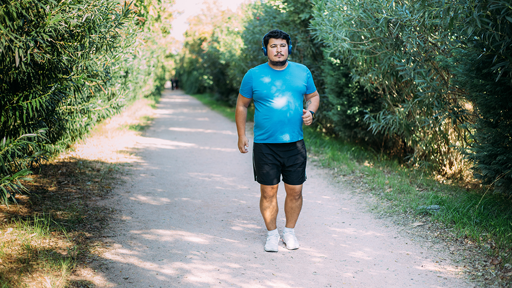 Latino man in blue shirt black shirt and over the ear headphones walks along a gravel trail