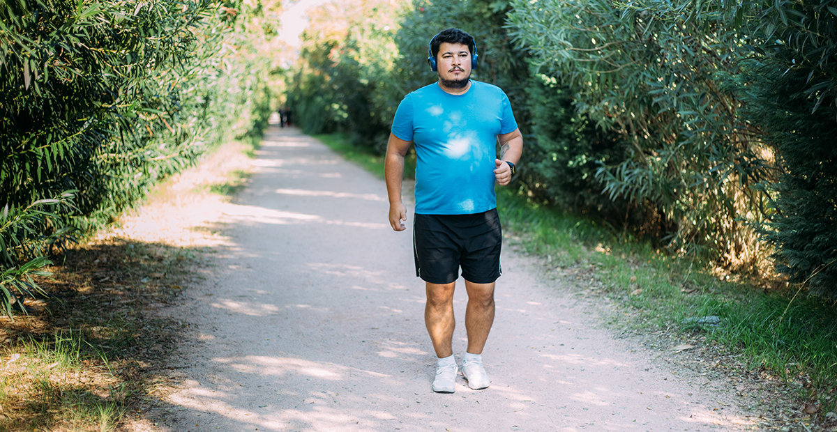 Latino man in blue shirt black shirt and over the ear headphones walks along a gravel trail