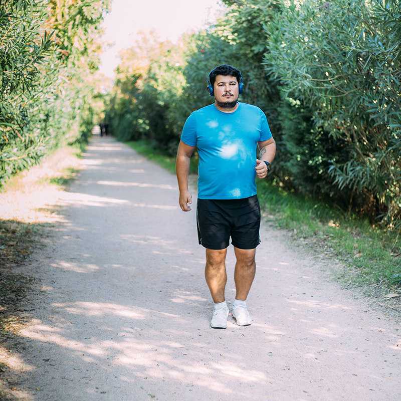 Latino man in blue shirt black shirt and over the ear headphones walks along a gravel trail