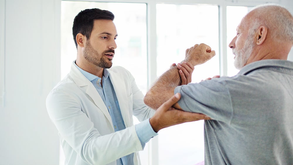 Closeup side view of an early 60's senior gentleman having some shoulder pain. He's at doctor's office having medical examination by a male doctor. The doctor is touching the sensitive area and trying to determine the cause of pain.