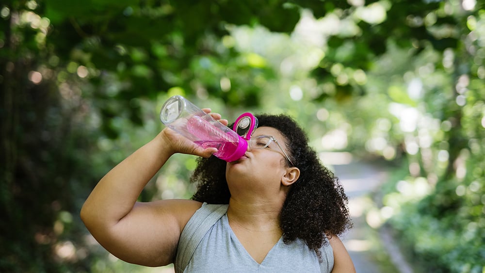Black woman with natural hair on nature trail drinking out of pink water bottle
