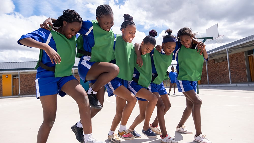 Group of teenage netball girls huddle together and celebrate scoring a goal in the match at their school