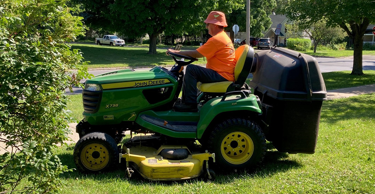 Man in orange shirt and orange hat driving green and yellow lawnmower on green grass