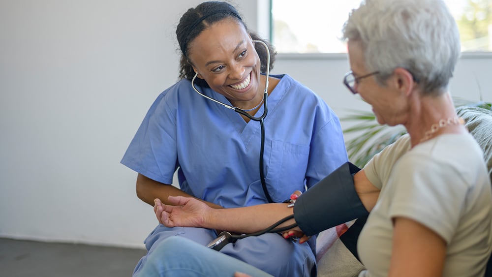 A female mixed race nurse of Hispanic and Latin descent sits on a sofa with her senior patient and checks the aging woman's blood pressure.