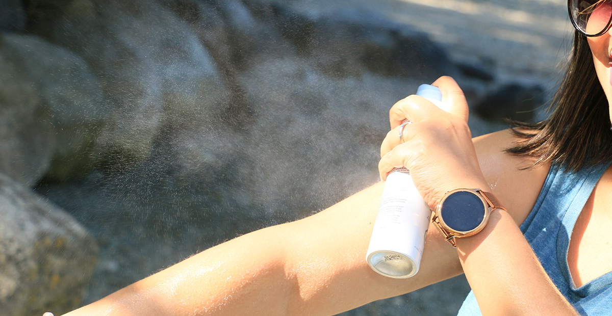 A woman spraying sunscreen on her bare arm. She is wearing sunglasses, a blue sleeveless top, a watch, bracelet, and holding an aerosol can of sunscreen