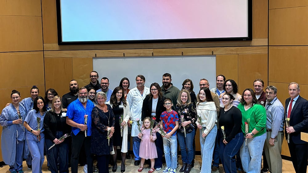 A group of healthcare workers stand in an auditorium with a patient whose life they saved