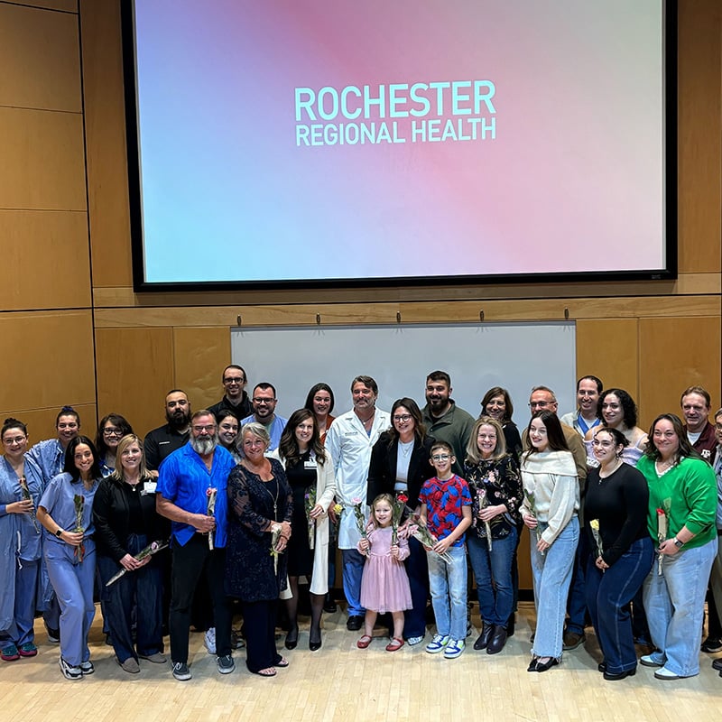 A group of healthcare workers stand in an auditorium with a patient whose life they saved