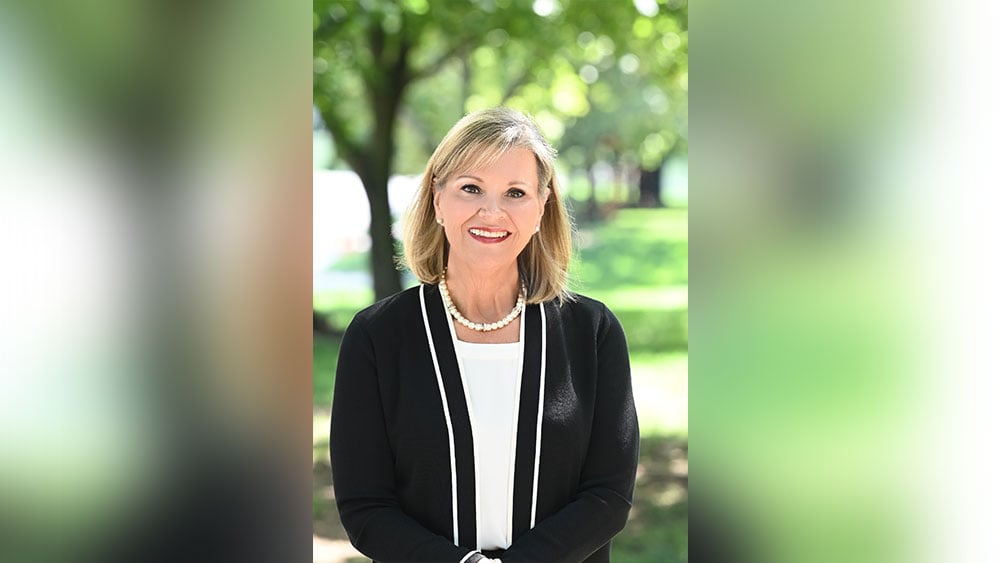 Tricia Gatlin is standing in front of green grass and trees. She is dressed in a business suit and smiling