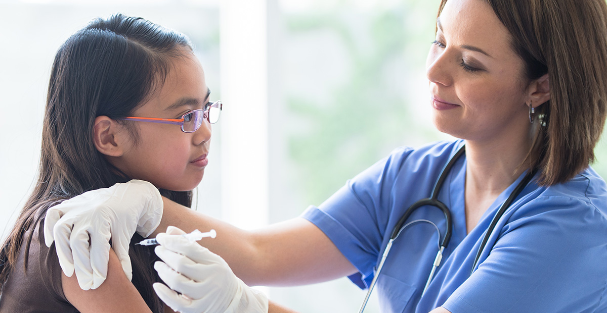 A young Asian girl sits up on an exam table with her sleeve over her shoulder, as a female Latin nurse gives the girl her immunization needle. The patient is dressed casually in a t-shirt and the nurse is wearing blue scrubs and white medical gloves for protection, as she holds the young girls shoulder with one hand and gives the injection with the other.