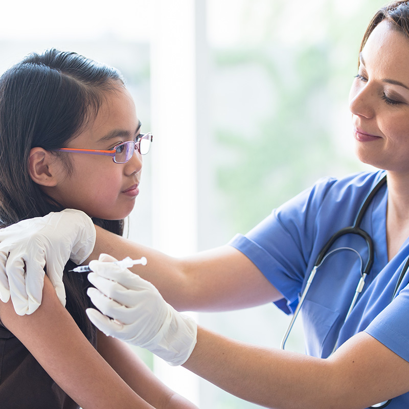 A young Asian girl sits up on an exam table with her sleeve over her shoulder, as a female Latin nurse gives the girl her immunization needle. The patient is dressed casually in a t-shirt and the nurse is wearing blue scrubs and white medical gloves for protection, as she holds the young girls shoulder with one hand and gives the injection with the other.