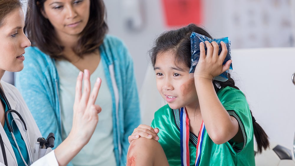 Serious female ER doctor holds up three fingers while talking with injured female elementary age soccer player with mom in the background