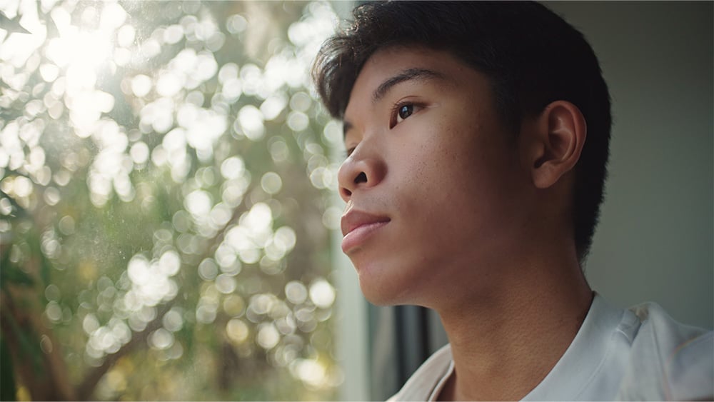 Asian teenage boy looking through the window with sunlight