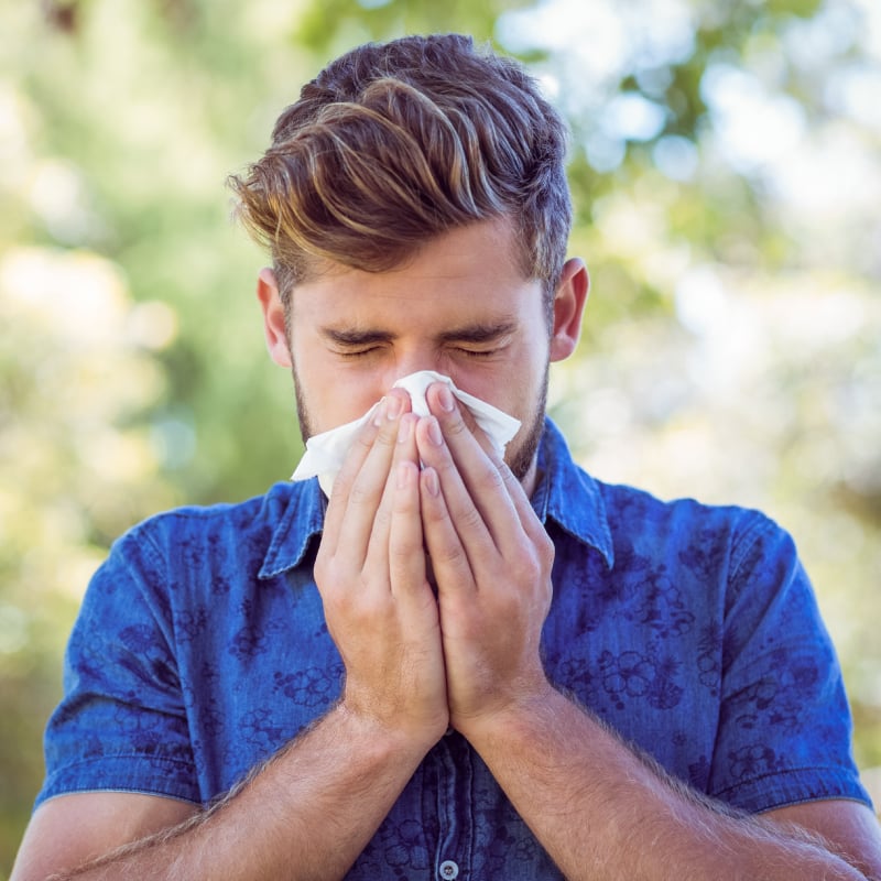A white man with styled hair and a blue polo blows his nose into a tissue outdoors