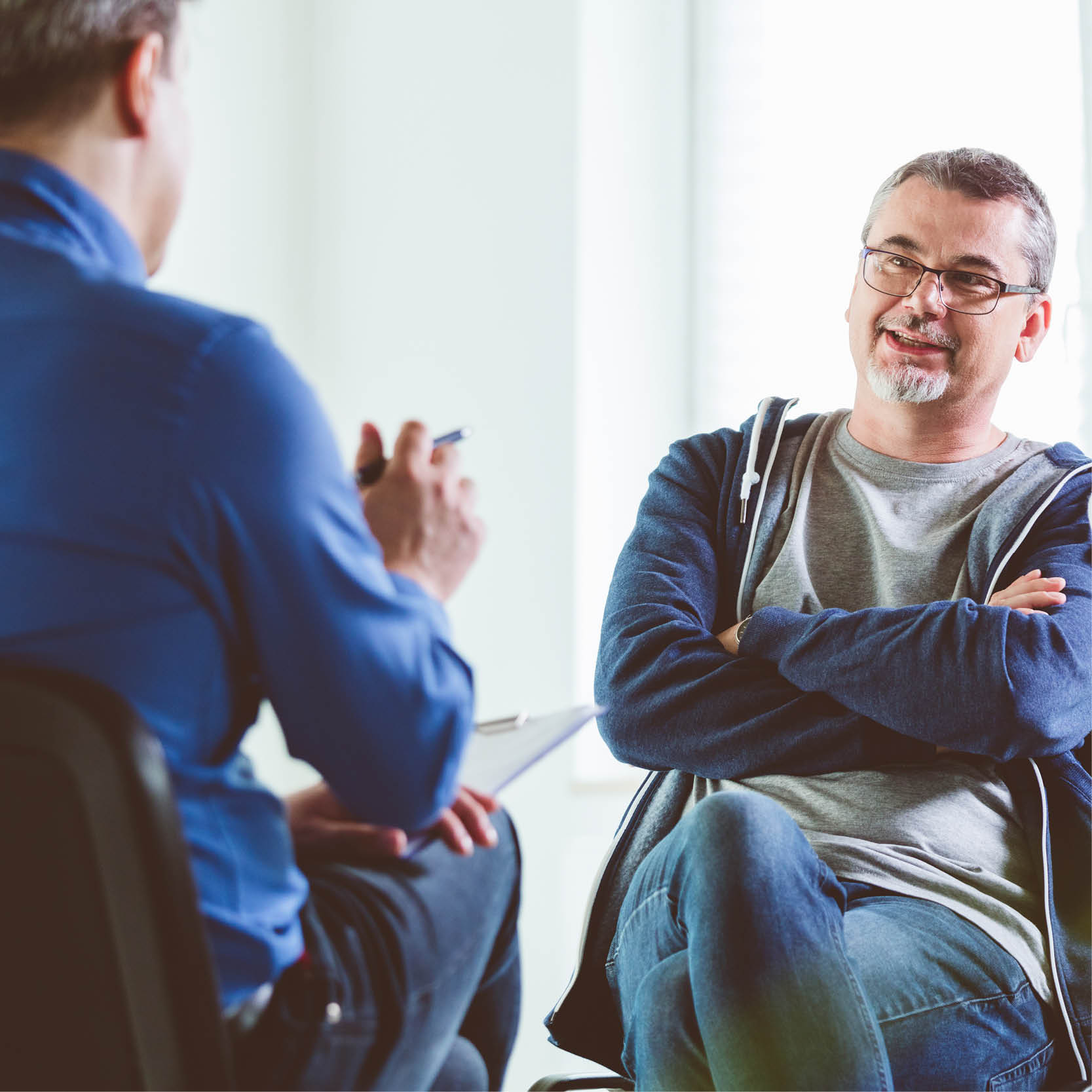 A middle-aged white man in a zip-up hooded sweatshirt and glasses listens to a healthcare provider talk in an office