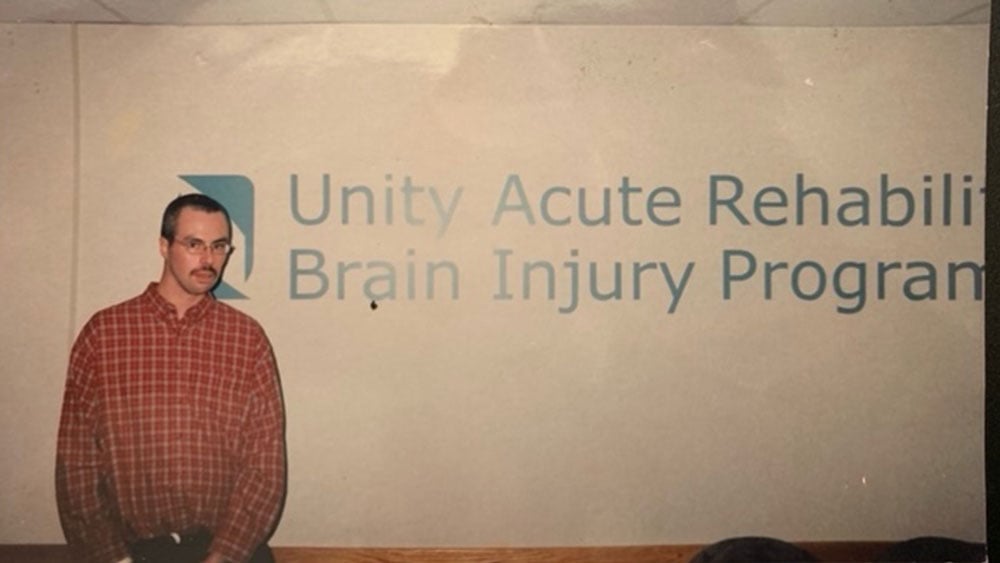 A white man with a mustache wearing a red and white checkered shirt standing in front of a white wall that says Unity Acute Rehabilitation Brain Injury Program in blue lettering