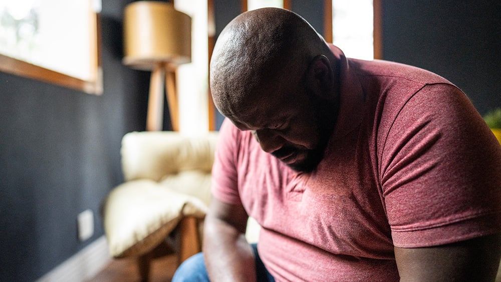 Sad middle aged Black in maroon t-shirt looking down while sitting in living room