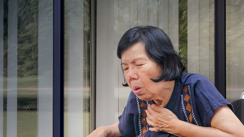 Older Asian woman coughing while next to window with vertical blinds