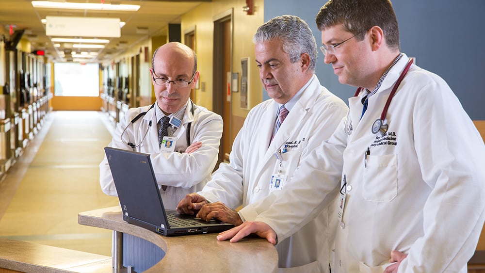 Three smiling white male doctors looking at laptop in hospital hallway