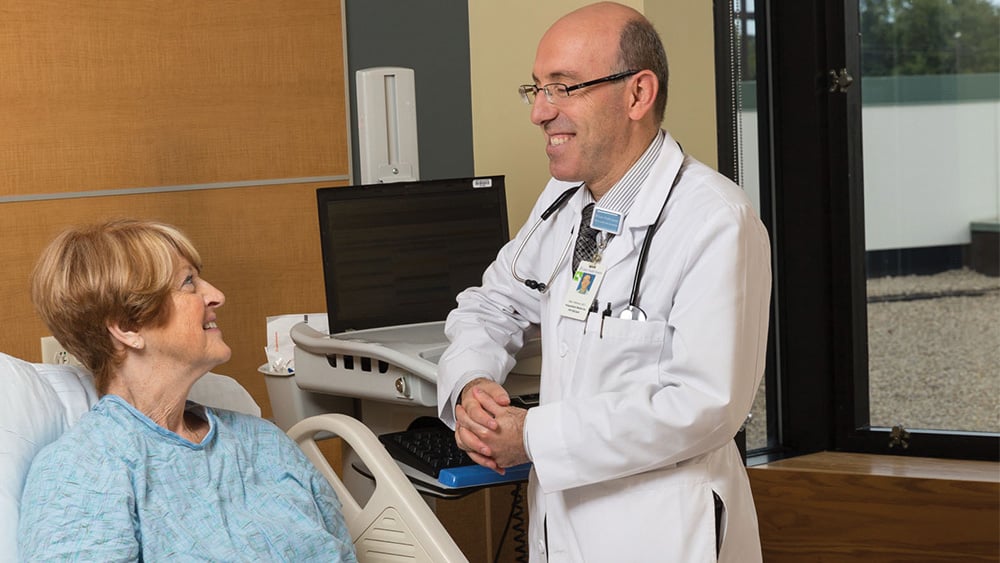 Older white female patient in hospital bed talks with balding white male doctor