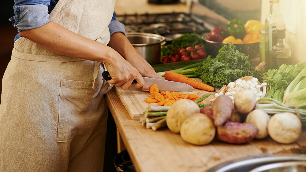 Person in apron chopping carrots at a kitchen counter, filled with vegetables.